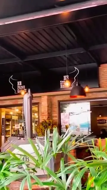 Salão com o logo HB Steak House na parede, luminárias pendentes e plantas