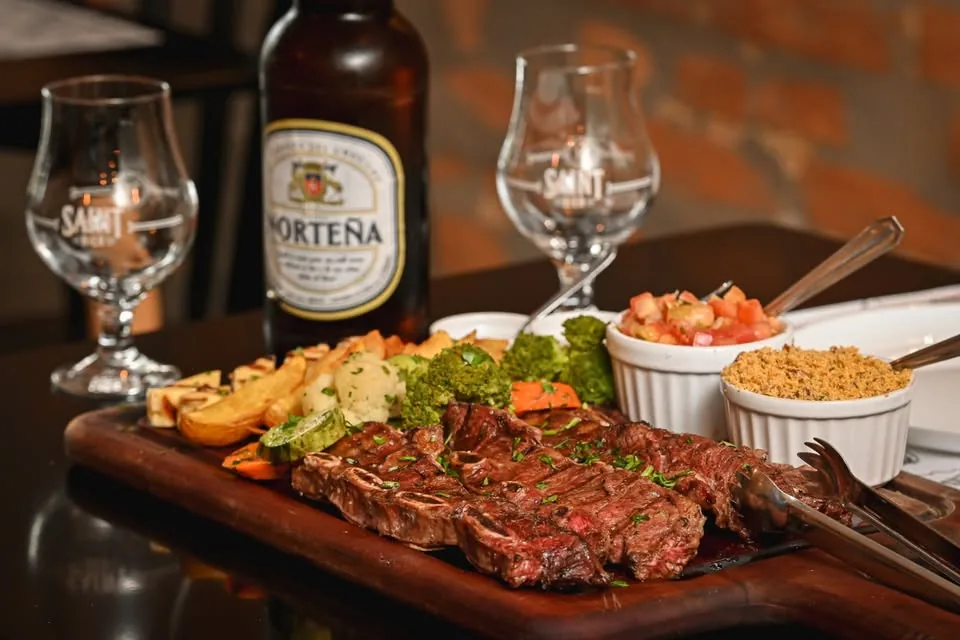 Tábua com carne grelhada fatiada, acompanhamentos em potes e cerveja